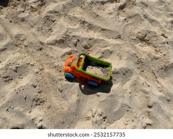 Orange and green toy truck on sandy beach, partially filled with sand, symbolizing fun and creative playtime outdoors. - Powered by Shutterstock