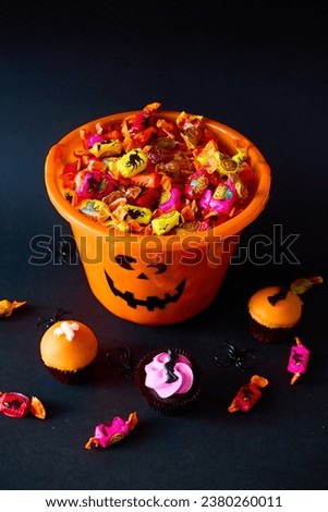 An orange halloween bucket full of coulourful candies on the black background, decorated with cupcakes