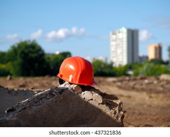 Orange Helmet On A Background Of A Vacant Lot. A Lot Of Sun. In The Distance, City, High-rise Buildings, Trees. Wasteland - A Place Under Construction