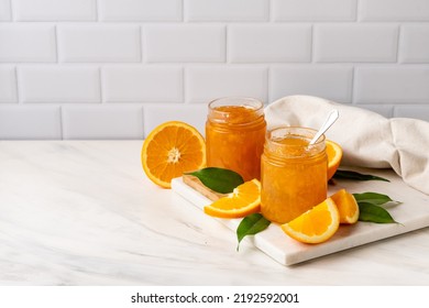 Orange jam in a glass jar and fresh oranges on a white marble background