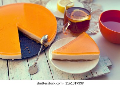 Orange Jelly Cheesecake (cheese Cake) On White Wooden Background. Piece Of Jelly Mousse Cheesecake Sliced Of Whole Pie Tart, Lemon Tea Cup. Homemade Mascarpone Cheesecake With Orange Fruit Gelatin