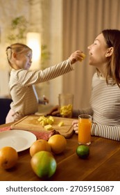 Orange juice on table while daughter feeds fruit to smiling mother in warm playful mood. Concept of drink marketing, family lifestyle visuals and emotional healthy routine.