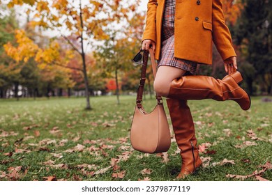 Orange knee high boots. Fashionable woman wearing stylish blazer plaid mini skirt walking in fall park with beige handbag among leaves. Copy space
