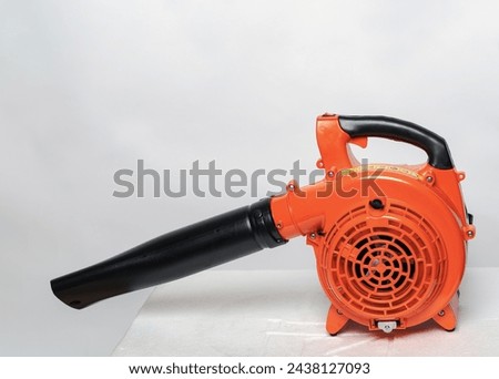 Orange leaf blower with a black nozzle. Its positioned on a light surface against a grey backdrop. It appears sturdy and designed for outdoor cleaning tasks, showcasing its utility and efficiency.
