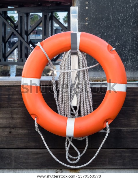 Orange life preserver or life buoy with reflective\
silver stripes, rope handles and attached coiled rope on hook\
located on city dock near\
bridge