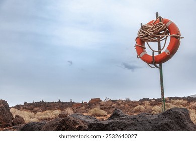 Orange lifebuoy is attached to a metal pole in a deserted volcanic landscape under a cloudy sky - Powered by Shutterstock