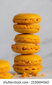 Orange Macaroons Piled On Top Of Each Other With Macaroon Crumbs In The Foreground And An Out Of Focus Macaroon In The Bottom Left Hand Corner Of The Foreground. White Marble Background And Foreground