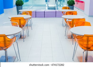 Orange Plastic Chairs And A White Table On Food Court In Mall With Purple Bench.