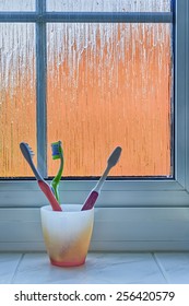 Orange plastic cup holding three toothbrushes (orange, green and pink) sitting on a bathroom windowsill against a frosted window, with distorted orange exterior [portrait format].