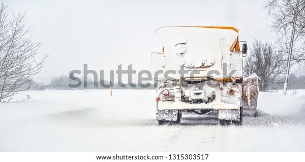Orange plough truck on snow covered road,\
gray sky and trees in background, view from back - winter road\
maintenance