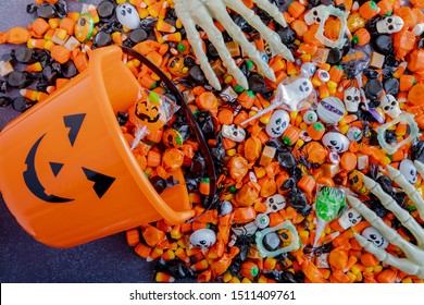 Orange pumpkin pail spilling Halloween candy on black stone surface with skeleton hands grabbing candy