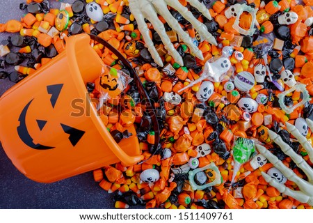 Orange pumpkin pail spilling Halloween candy on black stone surface with skeleton hands grabbing candy