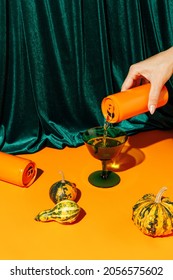 Orange pumpkins and a woman's hand pouring a beverage drink from an orange can against plush velvet curtain background. Creative Halloween and Thanksgiving concept. Contemporary fall still life idea.