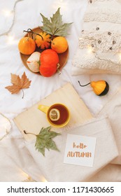 Orange Pumpkins, Wooden Tray, Cup Of Tea, Dry Leaves, Beige Plaid, Pillows, Glowing Garland Lights And Card With Text HELLO AUTUMN On Bed With White Linen. Home Cozy Fall Decor. Flat Lay, Top View.