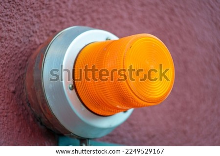 Orange saftey light on side of red building with stucco exterior and gray metal housing for bulb in an industrial area. In an urban area in the business or manufacturing districts for a warehouse.