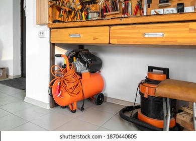 Orange small portable industrial power air compressor with coil hose and pneumatic gun and vacuum cleaner at home warehouse garage under wooden workbench. Electric tools and equipment