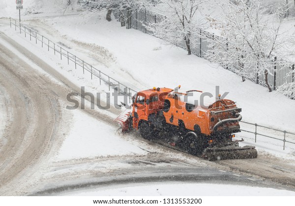 Orange snow plow truck or\
street sweeper machine cleans the road surface from snow. February\
snowfall
