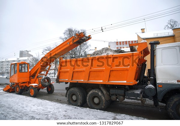 Orange snow removal machine loads snow into a\
dump truck for removal from city. Emergency cleans road after heavy\
snowfall  by municipal workers. Road cleaning with special\
equipment vehicle