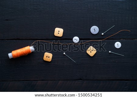 Orange spool of thread, pins and buttons for sewing on a wooden black background. 