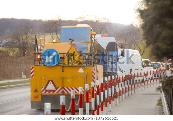 An orange street-cleaning machine stands in a\
traffic jam near the sidewalk designated by signal barriers. Road\
marking signs during road works. Restriction of traffic. Mechanized\
street cleaning