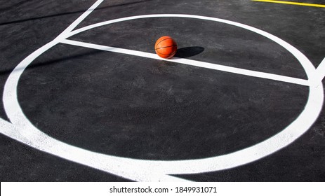 Orange Striped Basketball Ball Stands In Circle On The Marking Playing Field With Asphalt Tarmac, Sports Object Lit By Sun Light On Playground With Copy Space, Nobody.