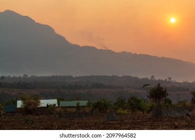 Orange Sun In Orange Sky At Sunset In A Haze Over A Hill And A Local House With A Field At A Coffee Farm. Bolaven Plateau, Laos