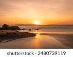 An orange sunrise with a diffused and colorful sky with offshore islands in the distance and a sandy tropical beach with rocks reflecting sunshine in the wet sand in Bowen in Queensland, Australia.