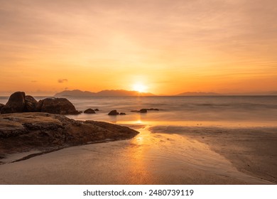 An orange sunrise with a diffused and colorful sky with offshore islands in the distance and a sandy tropical beach with rocks reflecting sunshine in the wet sand in Bowen in Queensland, Australia. - Powered by Shutterstock