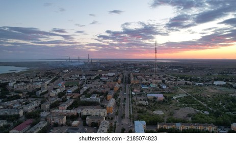 Orange Sunset Over A Small Town. Top View From A Drone. Blue-purple Clouds Against Background Of Houses, A Smoking Factory And A TV Tower. Cars Drive. There Is A Bright Light At The Metal Drain Plant