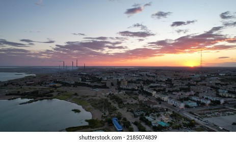 Orange Sunset Over A Small Town. Top View From A Drone. Blue-purple Clouds Against Background Of Houses, A Smoking Factory And A TV Tower. Cars Drive. There Is A Bright Light At The Metal Drain Plant