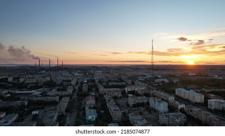 Orange Sunset Over A Small Town. Top View From A Drone. Blue-purple Clouds Against Background Of Houses, A Smoking Factory And A TV Tower. Cars Drive. There Is A Bright Light At The Metal Drain Plant