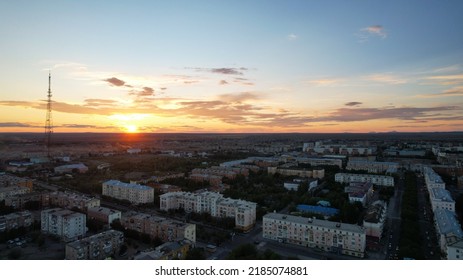 Orange Sunset Over A Small Town. Top View From A Drone. The Sun Is Sinking Below The Horizon. Blue-orange Clouds In The Sky. High TV Tower And Low Houses. Mountains Are Visible In Distance. Balkhash