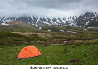 Orange tent with view to mountain range among low rainy clouds in overcast. Dramatic alpine landscape with snowy mountains in gray low clouds. Bleak atmospheric scenery of tundra under lead gray sky. - Shutterstock ID 2107883057