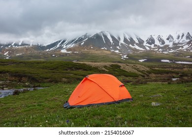 Orange tent with view to mountain range among low rainy clouds in overcast. Dramatic alpine landscape with snowy mountains in gray low clouds. Bleak atmospheric scenery of tundra under lead gray sky.