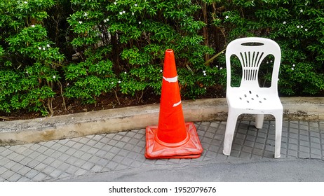 Orange Traffic Cone And White Plastic Chair Are On Footpath With White Flower And Green Plant Or Tree Background At Garden With Copy Space. Seat With Traffic Sing Object.