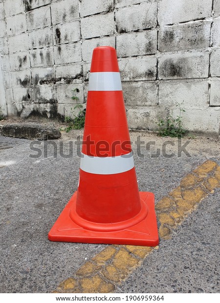 Orange traffic cone (With white and orange\
stripes) placed on the gray asphalt road in the parking lot with a\
yellow line beside the brick wall. Placed to prevent cars from\
parking this area.\
