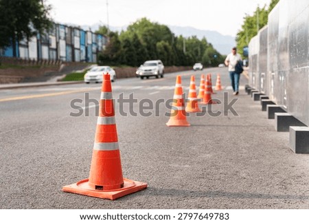 Orange traffic cones marking roadside safety zone on urban street with moving vehicles and pedestrian.