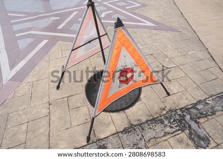 Orange triangle warning signs placed over an open manhole on a paved walkway for pedestrian safety.