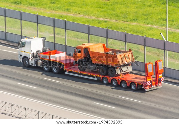 Orange truck dump with a load of\
soil in the body, transported on a trailer to the loading platform\
of another truck, help is broken breakdown\
evacuation
