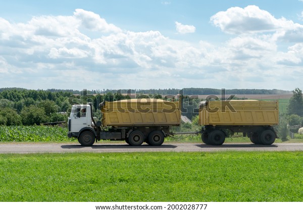An orange\
truck with a trailer transports grain along the highway on a\
beautiful natural landscape. The time of harvesting of grain crops\
and other sowing and harvesting\
operations