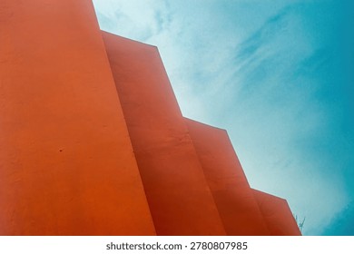 Orange wall forms a geometric composition against a vibrant blue sky, resembling an abstract stair pattern in urban architecture