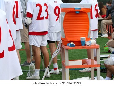 Orange Water Cooler On A Stand By The Athletes On The Bench Of A Lacrosse Game With Paper Cups.