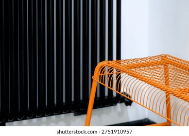 An orange wire table is positioned next to a black vertical radiator in a contemporary interior space with white walls and minimal decor