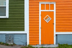 An Orange Wooden Exterior Door With A Diamond Shaped Window In A Building With Orange And Green Wood Clapboard. The Trim On The Building Is White In Colour. The Concrete Foundation Is Grey In Colour.