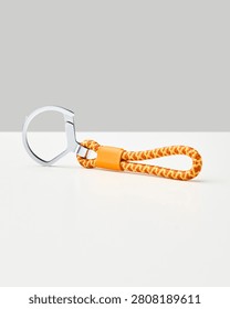 An orange and yellow patterned rope keychain with a silver metal ring rests on a white surface against a neutral light grey background, studio.