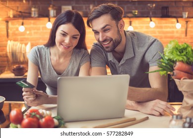 Order Food Online. Young Beautiful Couple In Kitchen Ordering Food Online From Restaurant. Family Of Two Using Laptop. Young Woman Paying With Credit Card. Nice Loft Kitchen Interior With Light Bulbs