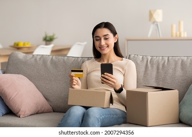 Order Goods, Delivery Of Purchases, Online Banking, Modern Shopping, Covid-19 Quarantine. Happy Young Arab Lady With Cardboard Boxes On Sofa In Living Room, Paying Bill With Credit Card And Phone