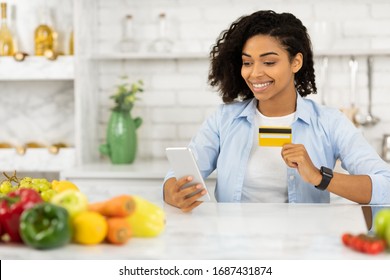 Ordering Food Online. Black Woman With Smart Phone And Credit Card Purchasing Grocery Delivery From Internet In Kitchen