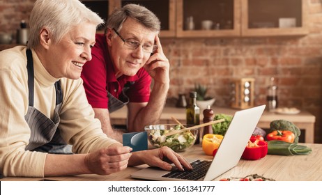 Ordering Food Online. Smiling elderly couple with laptop and credit card purchasing grocery delivery from internet while cooking together in kitchen