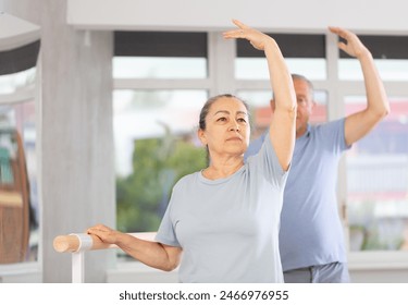 Ordinary elderly people stand in third position near ballet barre during group training in a dance studio - Powered by Shutterstock